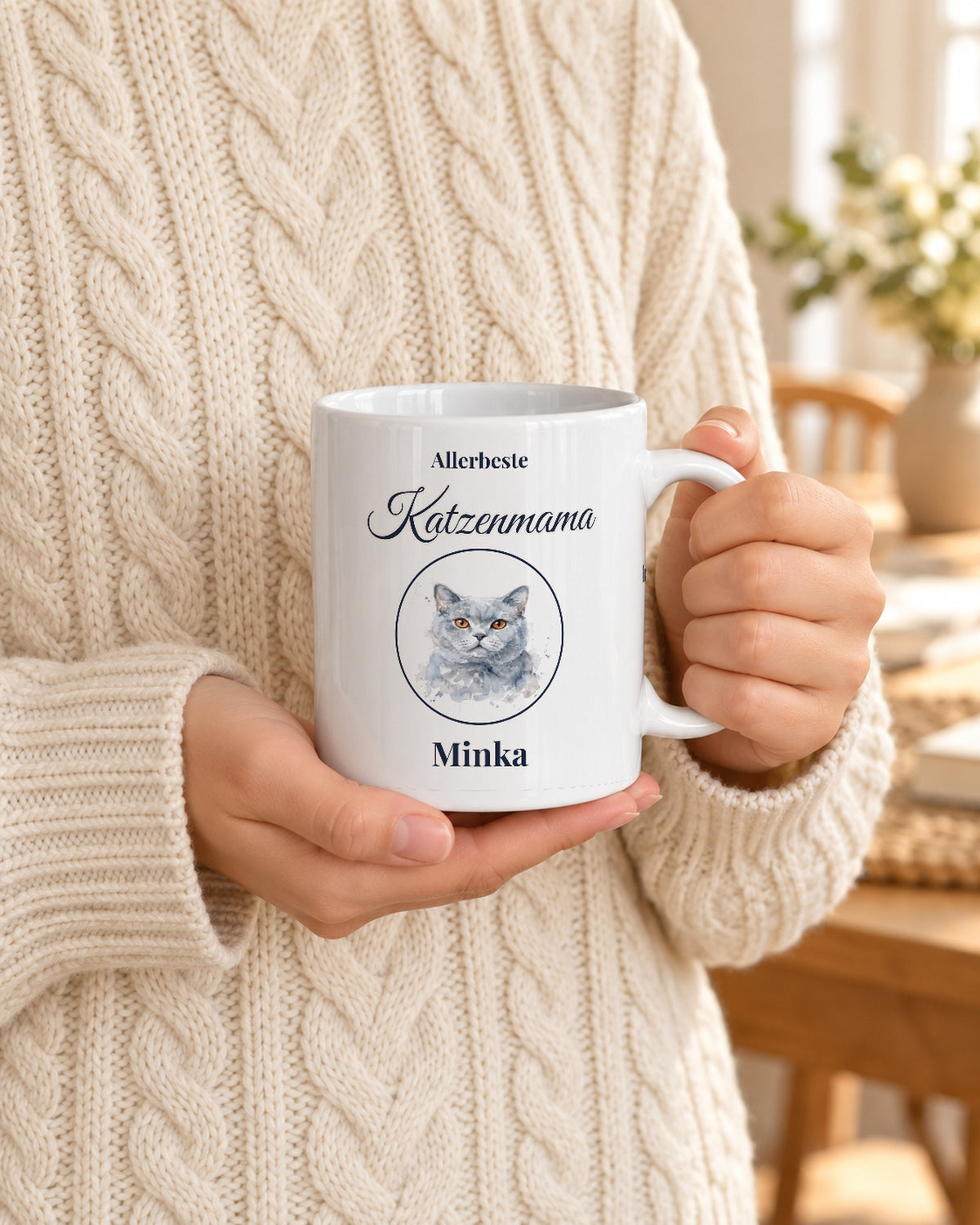 Weiße Tasse mit Katzenportrait im Aquarell-Stil, „Allerbeste Katzenmama“ und Namen, gehalten von Händen im Strickpulli