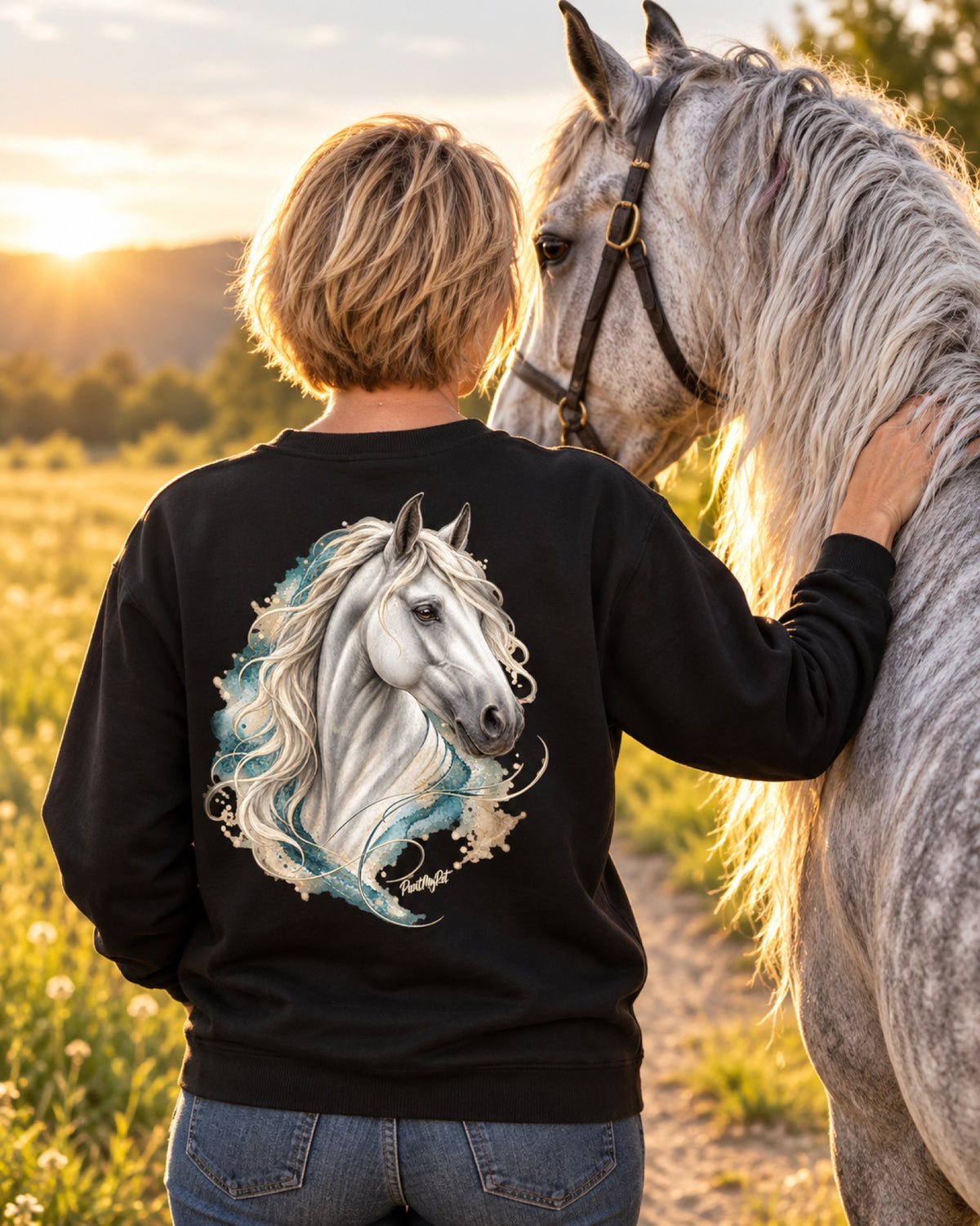 Frau im schwarzen Sweatshirt mit Schimmel im Aquarell-Stil auf dem Rücken, neben ihr ihr Pferd im Abendlicht