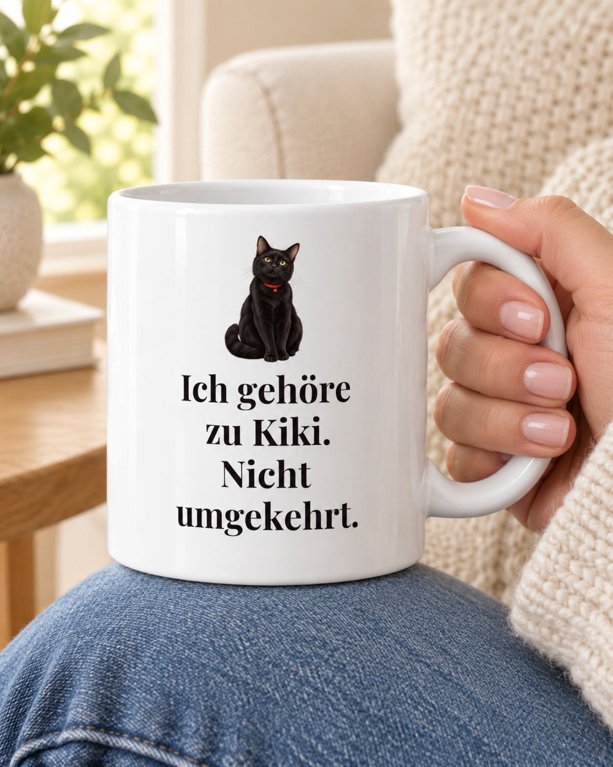 Weiße Katzentasse mit schwarzer Katze und Spruch Ich gehöre zu Kiki. Nicht umgekehrt., in der Hand auf dem Knie